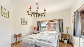A bedroom with a double bed, a chandelier, a window with curtains, a bedside table with a lamp, and a framed picture on the wall. - Le Molinel - vakantiewoning in hartje Koksijde (Koksijde)