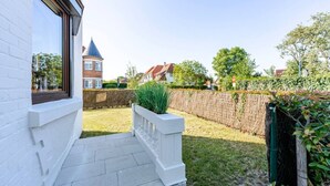 A residential property with a white exterior, a visible window, a paved entrance, a grassy yard, a wooden fence, and a distant building with a turret. - Le Molinel - vakantiewoning in hartje Koksijde (Koksijde)