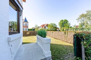 Ein Wohnhaus mit weißer Fassade, einem Fenster, einem gepflasterten Eingangsbereich, einem Rasen, einem Holzzaun und einem Gebäude mit einem Turm in der Ferne.