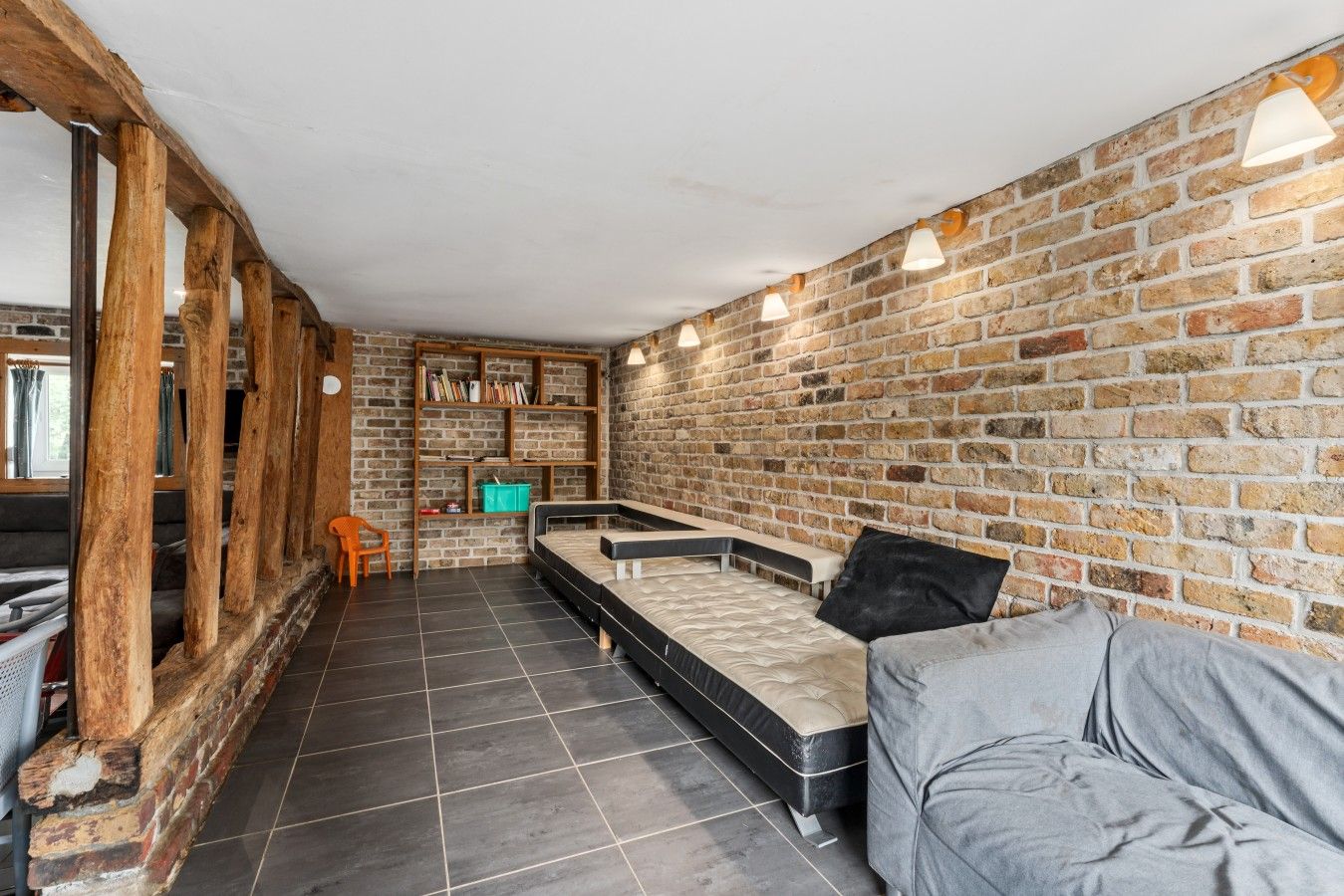 A room with a brick wall, a sofa, a bed, a wooden chair, and a bookshelf.