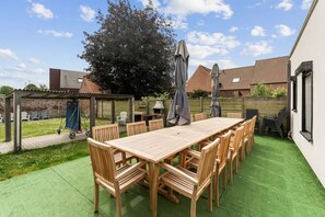 A patio with a wooden table and chairs, surrounded by a green lawn and a brick wall. - Barbara`s Guesthouse (Poperinge)