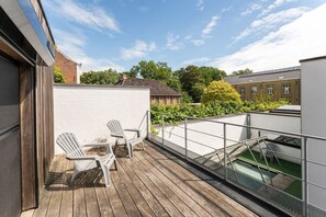 Eine Holzterrasse mit zwei weißen Stühlen, einem kleinen Tisch und einem Glasgeländer mit Blick auf eine Grünfläche.