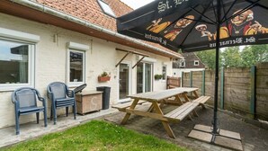 Area patio con tavolo e sedie di legno, un cestino e un ombrello nero con un logo.