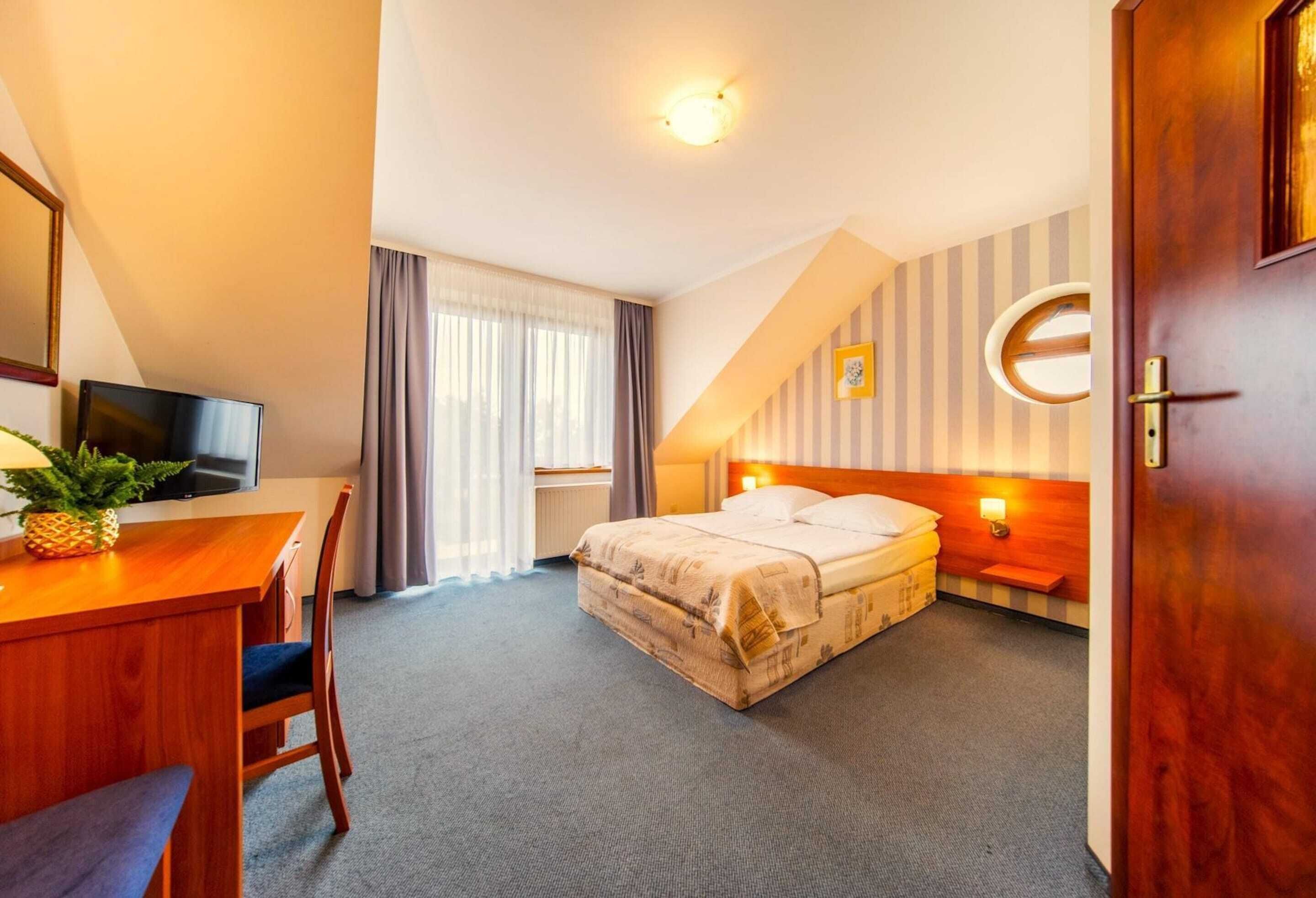 Une chambre d’hôtel avec un lit, un bureau et une chaise, une télévision et une fenêtre avec des rideaux.
