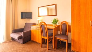 Quarto de hotel com sofá, escrivaninha com duas cadeiras, televisão, espelho e guarda-roupa.