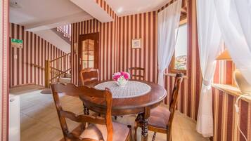 A dining area with a round wooden table, four chairs, and a vase of flowers. A staircase with striped wallpaper is visible in the background.