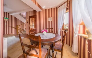 Un comedor con una mesa redonda de madera, cuatro sillas y un jarrón de flores. En el fondo, se aprecia una escalera con papel tapiz a rayas.