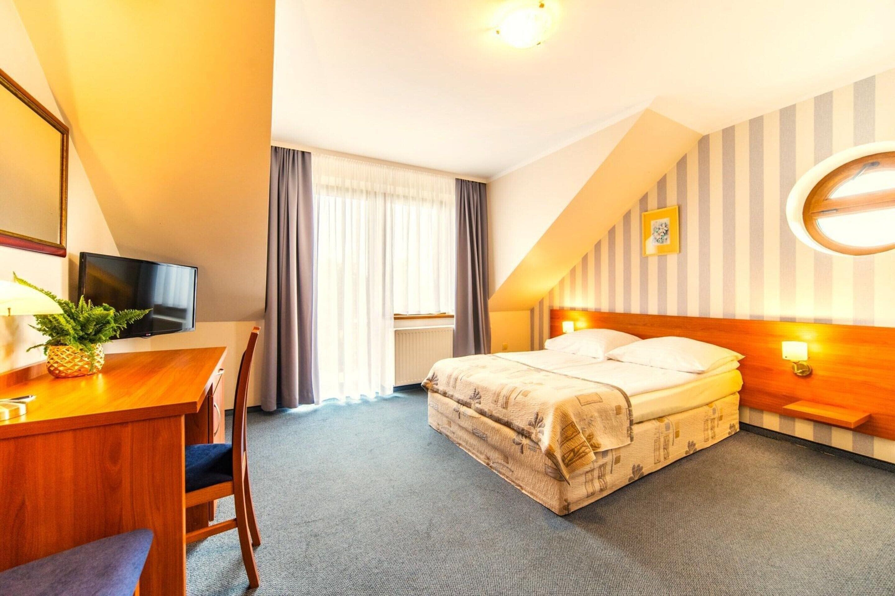 A hotel room with a bed, a desk with a television, a chair, a window with curtains, and a wall with a striped pattern.