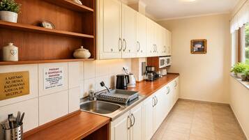 A kitchen with white cabinets, a wooden countertop, a sink, a coffee maker, and a kettle. There are signs on the wall and a plant on the window sill.