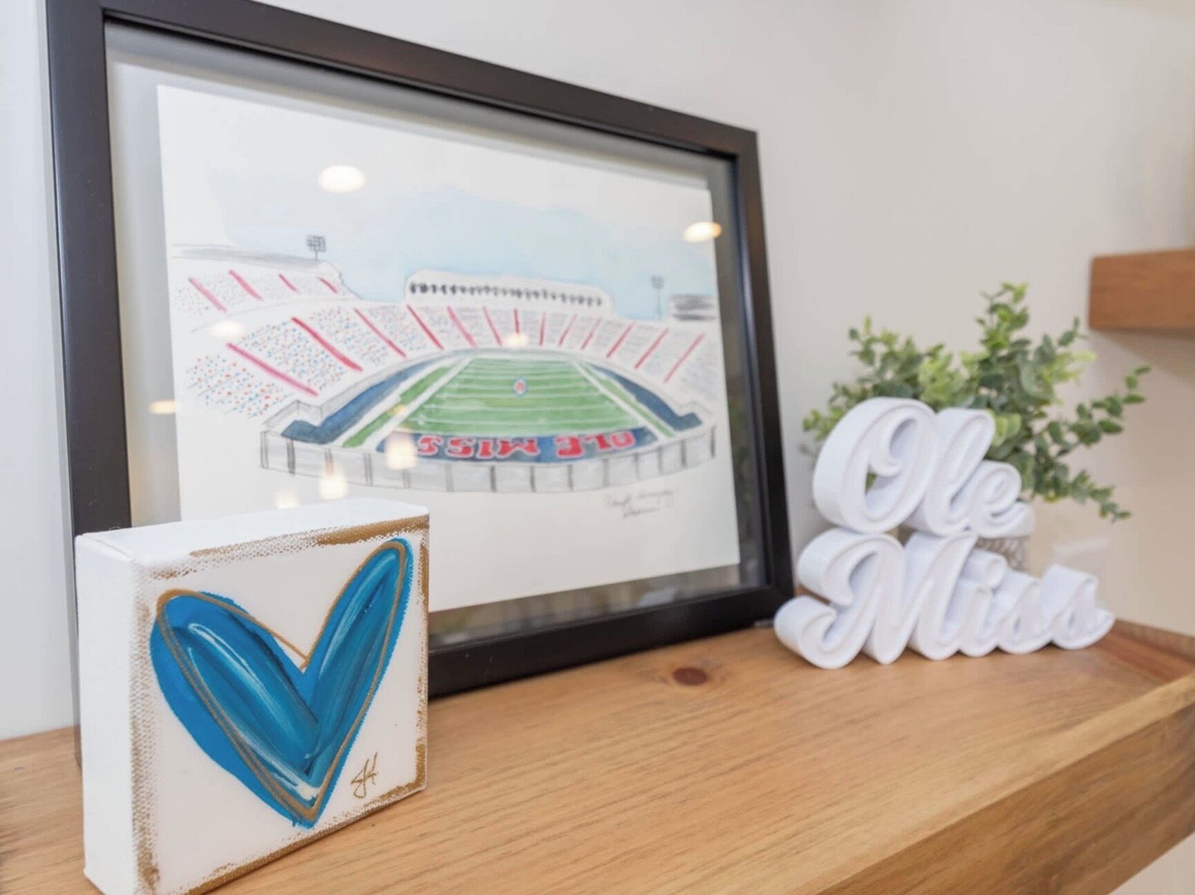 A framed drawing of a stadium with a heart-shaped object and a sign saying 'Ole Miss' on a wooden surface.