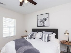 Une chambre à coucher avec un lit, un ventilateur de plafond, une fenêtre avec des stores, un tableau représentant un taureau accroché au mur et des lampes de chevet.