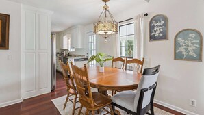 A dining room with a wooden table and chairs, a chandelier, and a view of the kitchen through a large window. - Stillwater Cottage - Dock + Fishing (Brunswick)