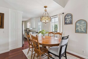 A dining room with a wooden table and chairs, a chandelier, and a view of the kitchen through a large window. - Stillwater Cottage - Dock + Fishing (Brunswick)