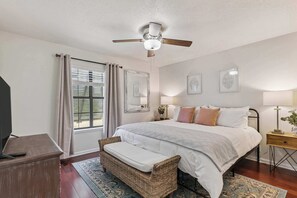 Ein Schlafzimmer mit einem Bett, Deckenventilator, einem Fenster mit Vorhängen, einem Nachttisch mit Lampe und einer kleinen Bank.