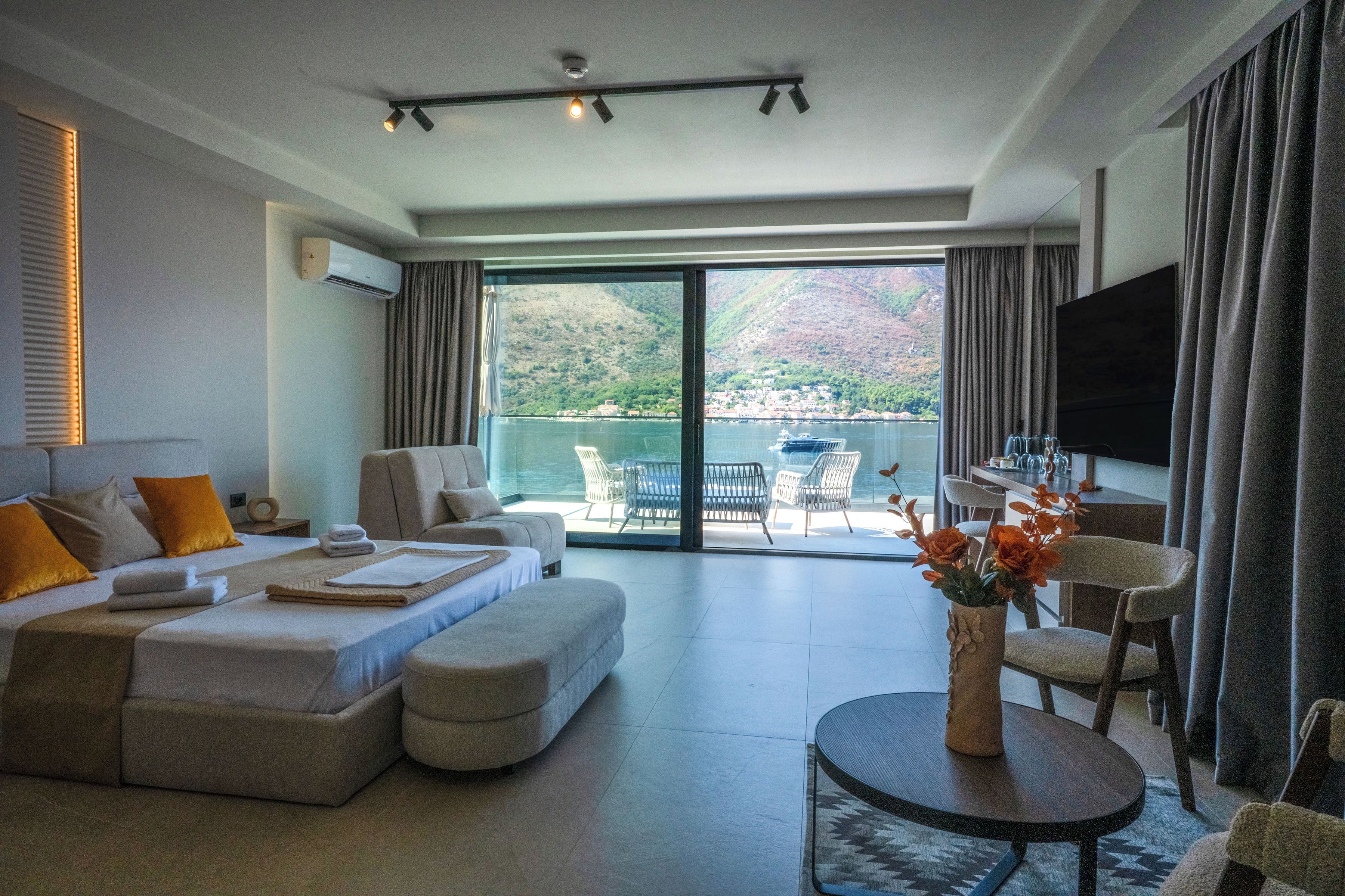 A modern hotel room with a large bed, a sofa, a dining area, and a balcony with a view of the mountains.