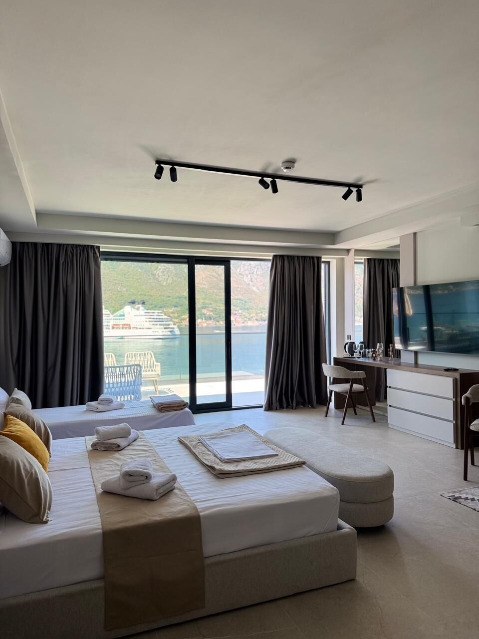 A modern hotel room with a bed, a desk, a TV, and a view of a cruise ship.