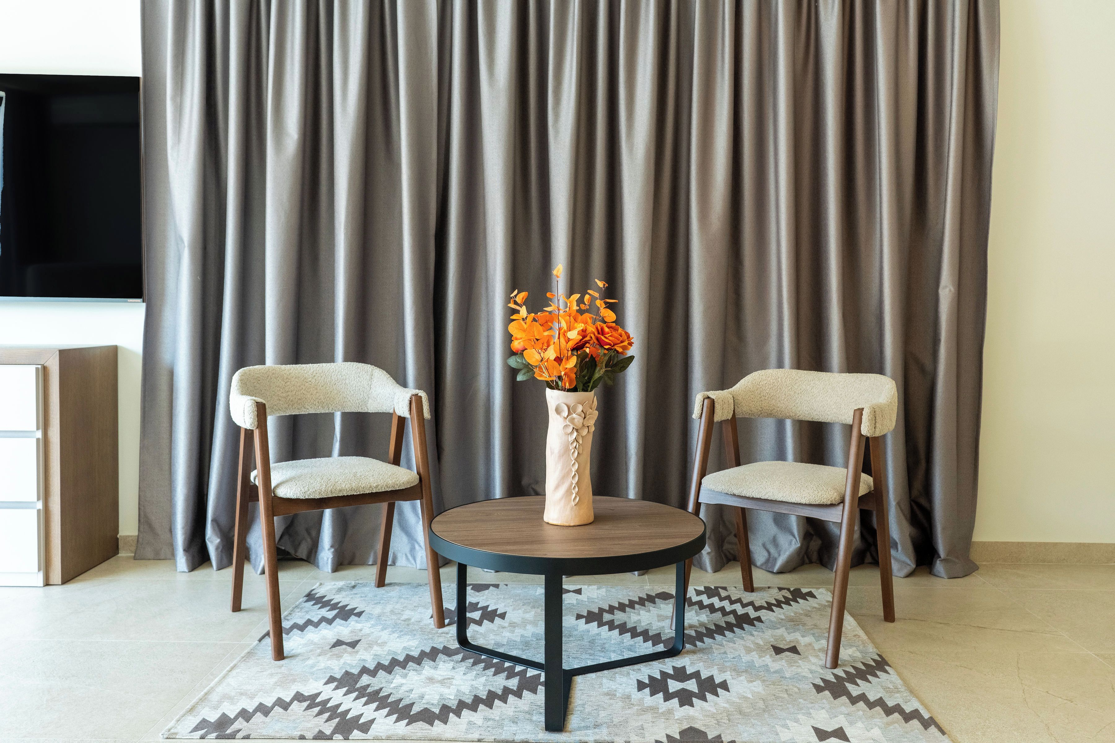 Ein Zimmer mit zwei Stühlen, einem kleinen Tisch, einer Blumenvase und einem Fernseher vor einem Vorhang.