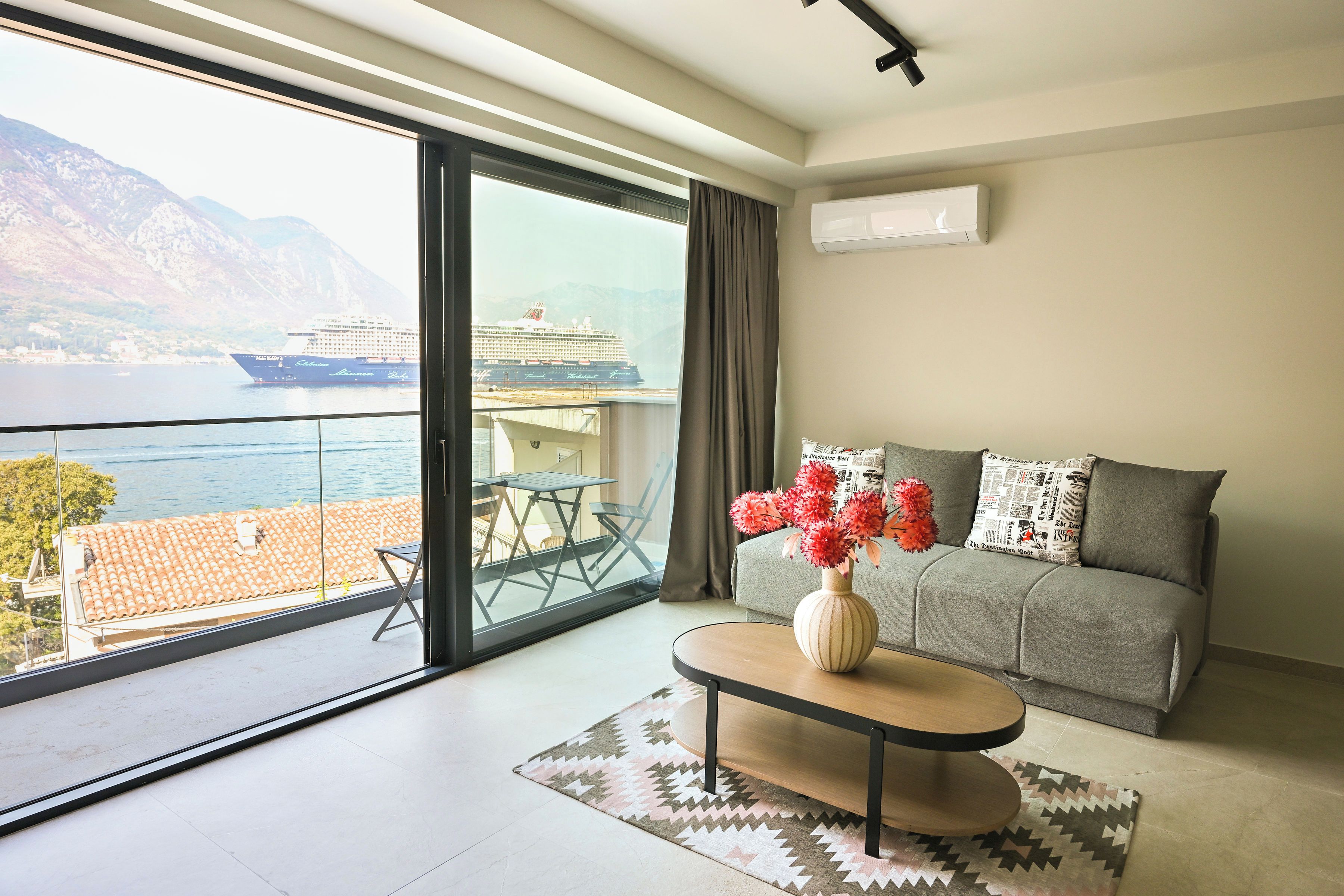 A modern living room with a large sliding glass door leading to a balcony, a sofa, a coffee table, and a vase with flowers.
