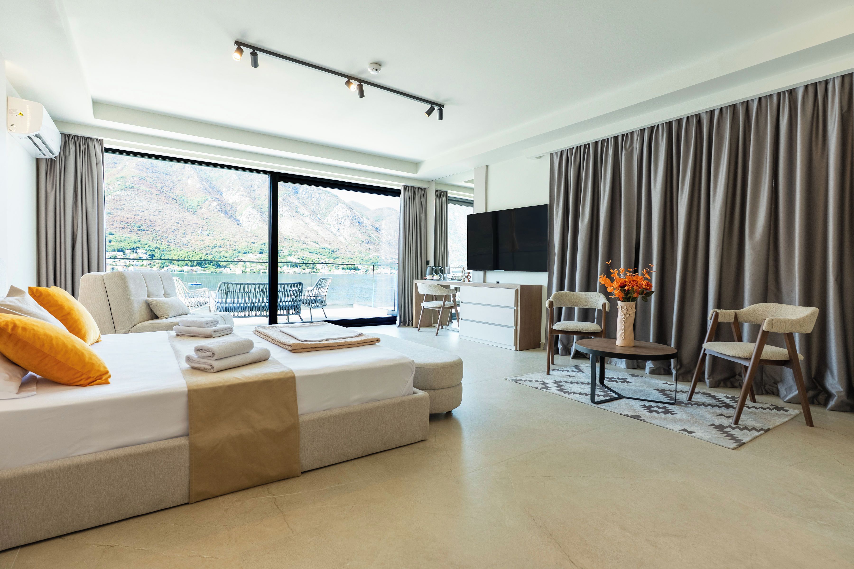 A modern hotel room with a large bed, a seating area with chairs, a flat-screen TV, and a view of mountains through the windows.