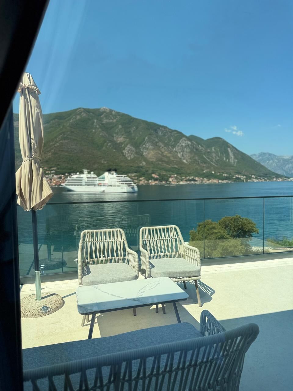 A balcony with wicker chairs and a table, overlooking a body of water with a cruise ship and mountains in the background.