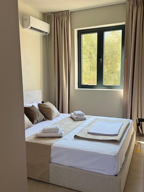 Una habitación de hotel con cama, ventana, cortinas, un equipo de aire acondicionado y una mesita de noche.