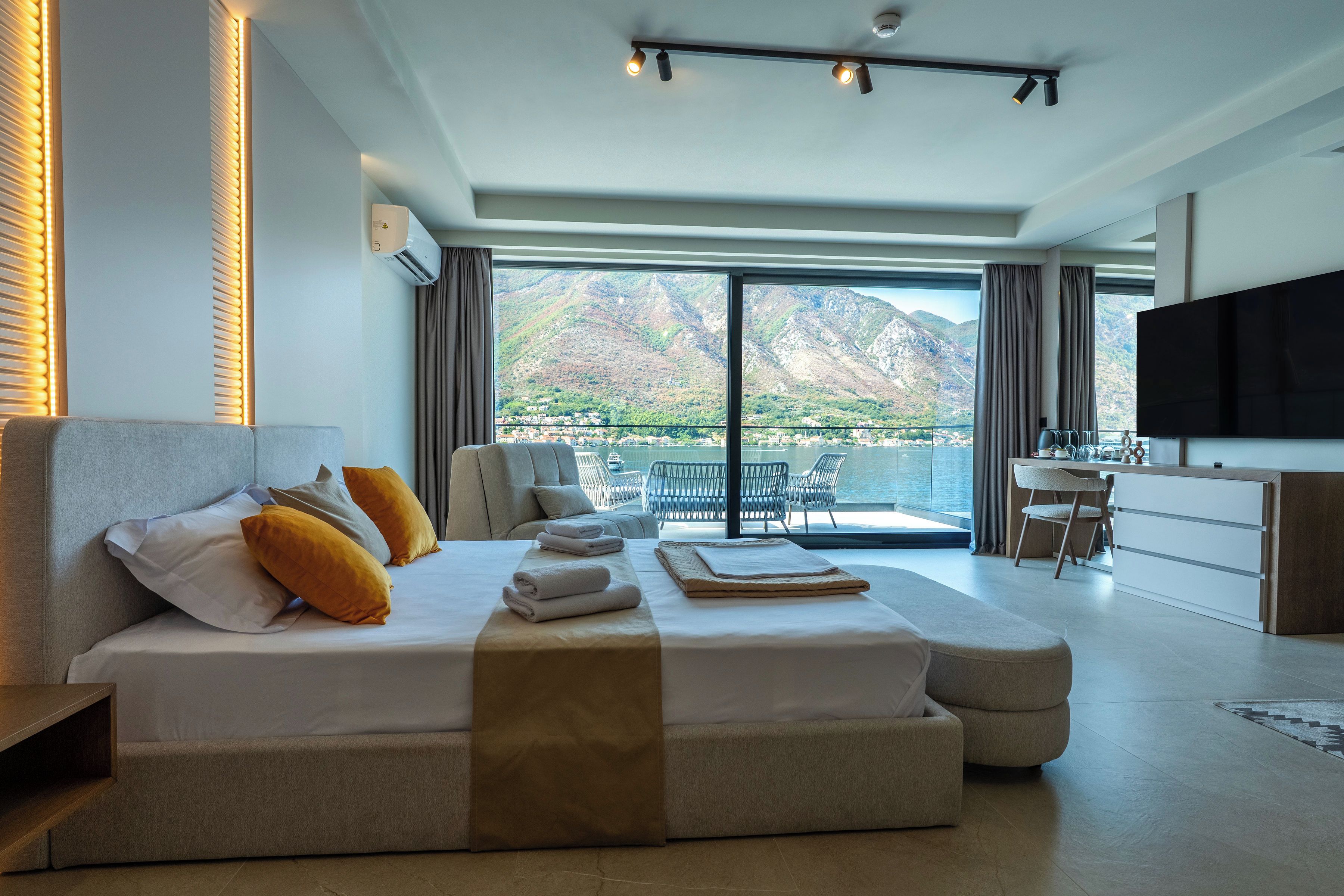 A modern hotel room with a large bed, a flat-screen TV, a balcony with a mountain view, and a kitchenette.