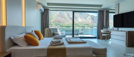 A modern hotel room with a large bed, a flat-screen TV, a balcony with a mountain view, and a kitchenette.