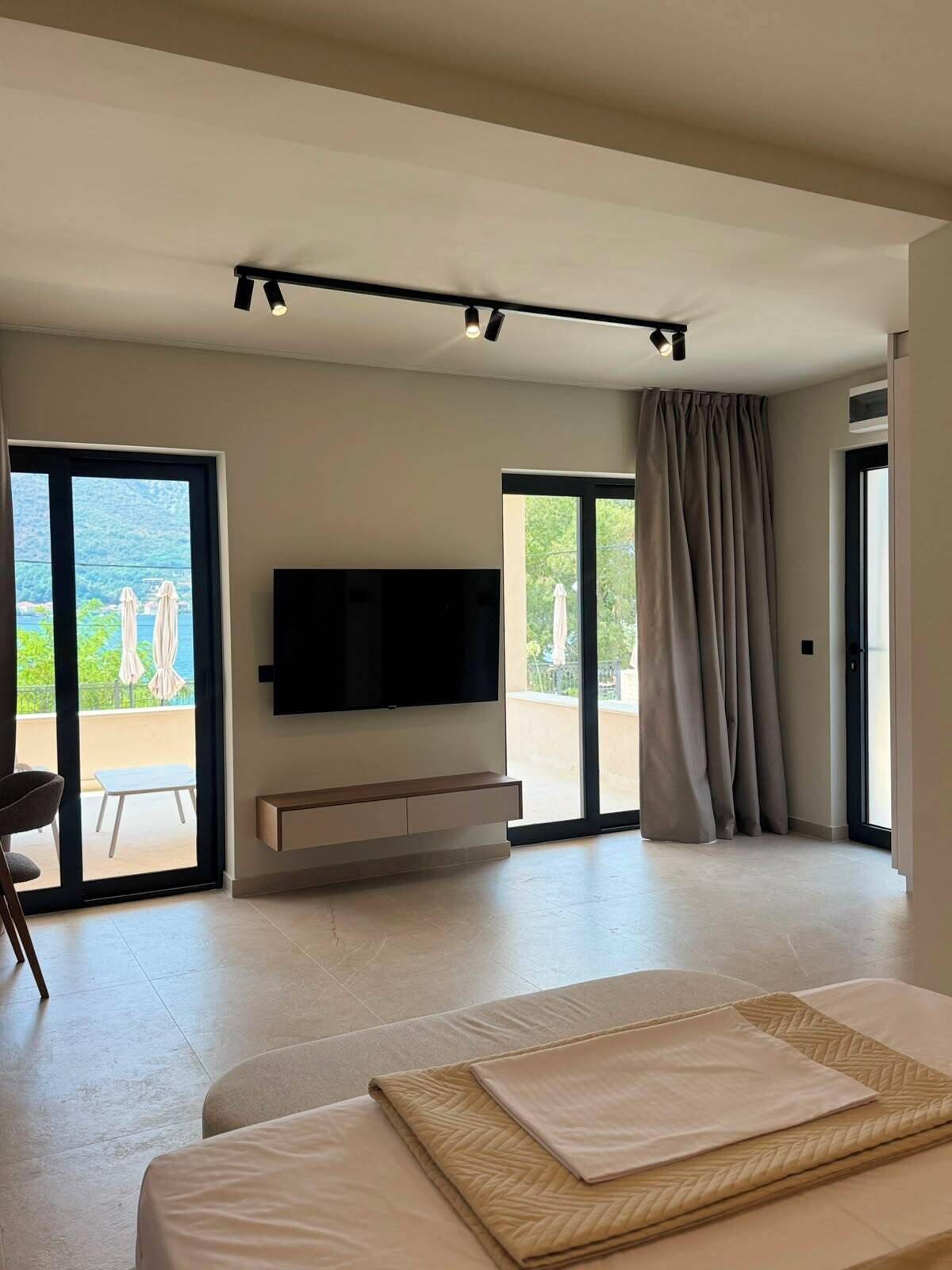 A modern bedroom with a large bed, a flat-screen TV mounted on the wall, and a sliding glass door leading to a balcony.