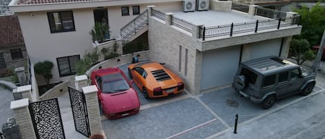 Una casa moderna de dos plantas con balcón, un camino de acceso con dos coches, y vistas a un cuerpo de agua y colinas al fondo.