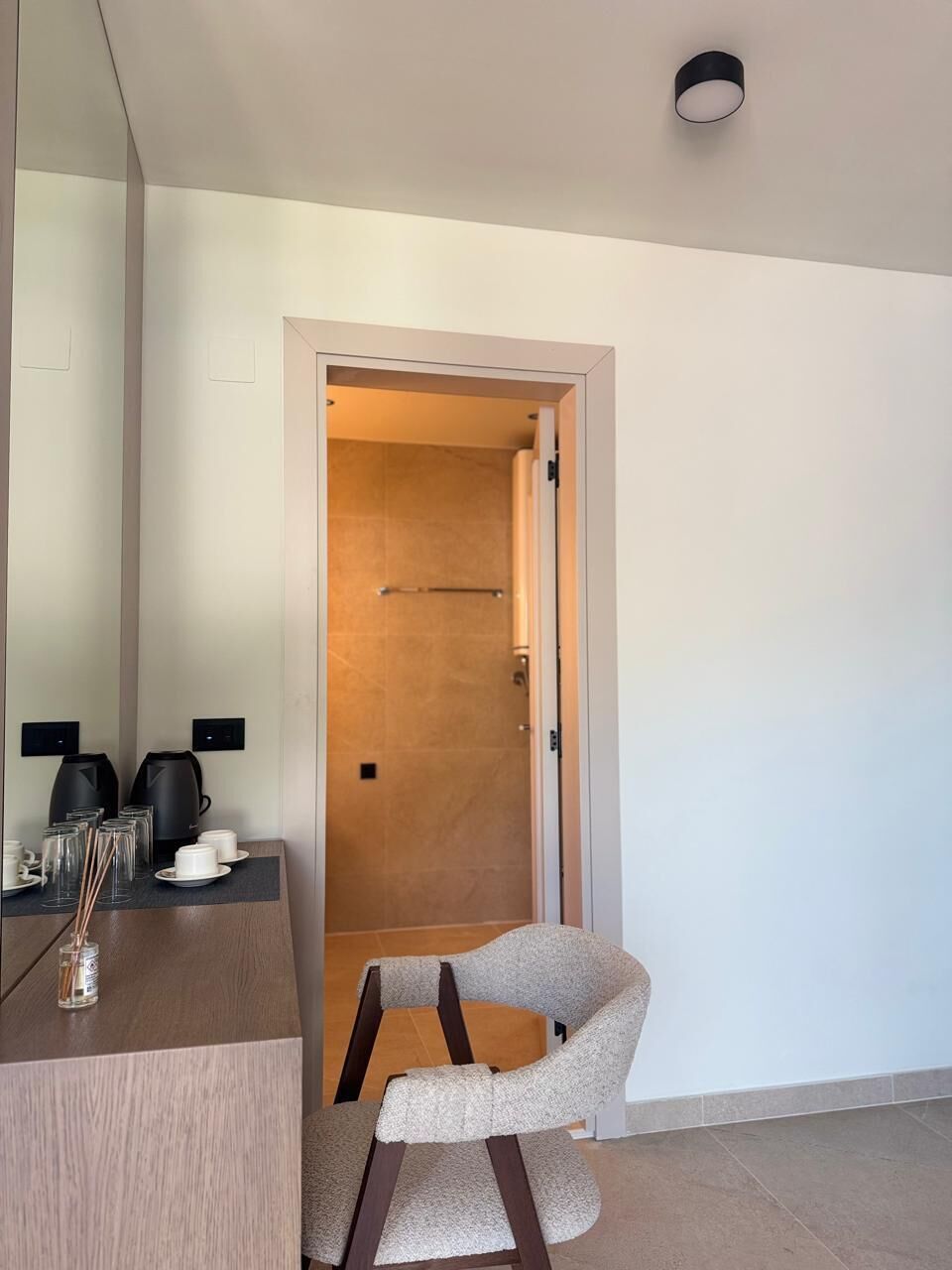 Une chambre d’hôtel moderne avec une porte en bois, un coin salon avec un fauteuil et une petite table sur laquelle se trouvent une machine à café et des verres.