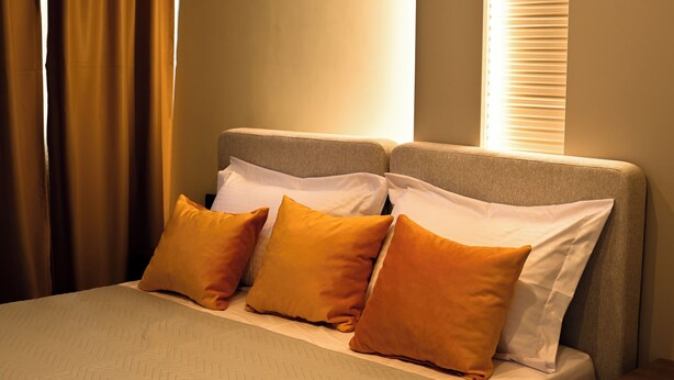 Habitación de hotel con cama, almohadas, mesita de noche y ventana con persianas.