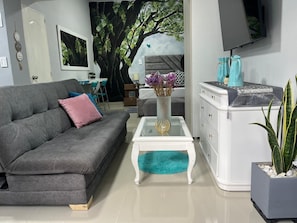 Ein modernes Wohnzimmer mit einem grauen Sofa, einem Glastisch, einer weißen Kommode und einem großen Wandgemälde mit einem Baum.