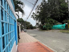 Eine Straßenszene mit Bürgersteig, einem blauen Gebäude mit Gitterstruktur und einer Straße, auf der sich rechts eine grüne Plane befindet.