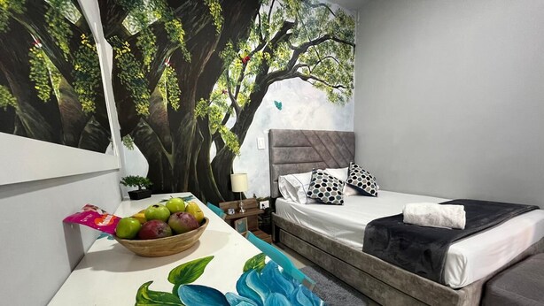 A bedroom with a bed, a nightstand, a wall mural, a table with a flower painting, and a fruit bowl. - Cozy House + Kitchen + AC + Pet-Friendly + TV + WiFi @ Cali (Cali)