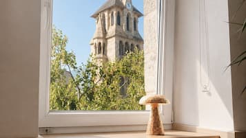 A room with a large window overlooking a historic building with a tower, a small wooden stool, and a plant.