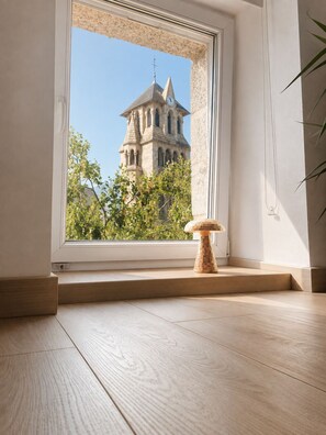 A room with a large window overlooking a historic building with a tower, a small wooden stool, and a plant.