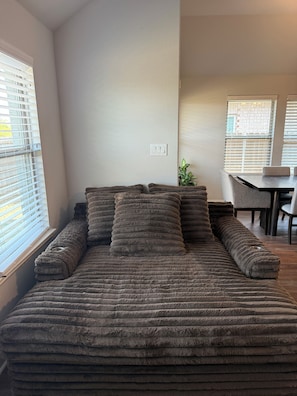 A room with a large, fluffy, multi-seater sofa against a wall, a dining area with a table and chairs, and a window with blinds.