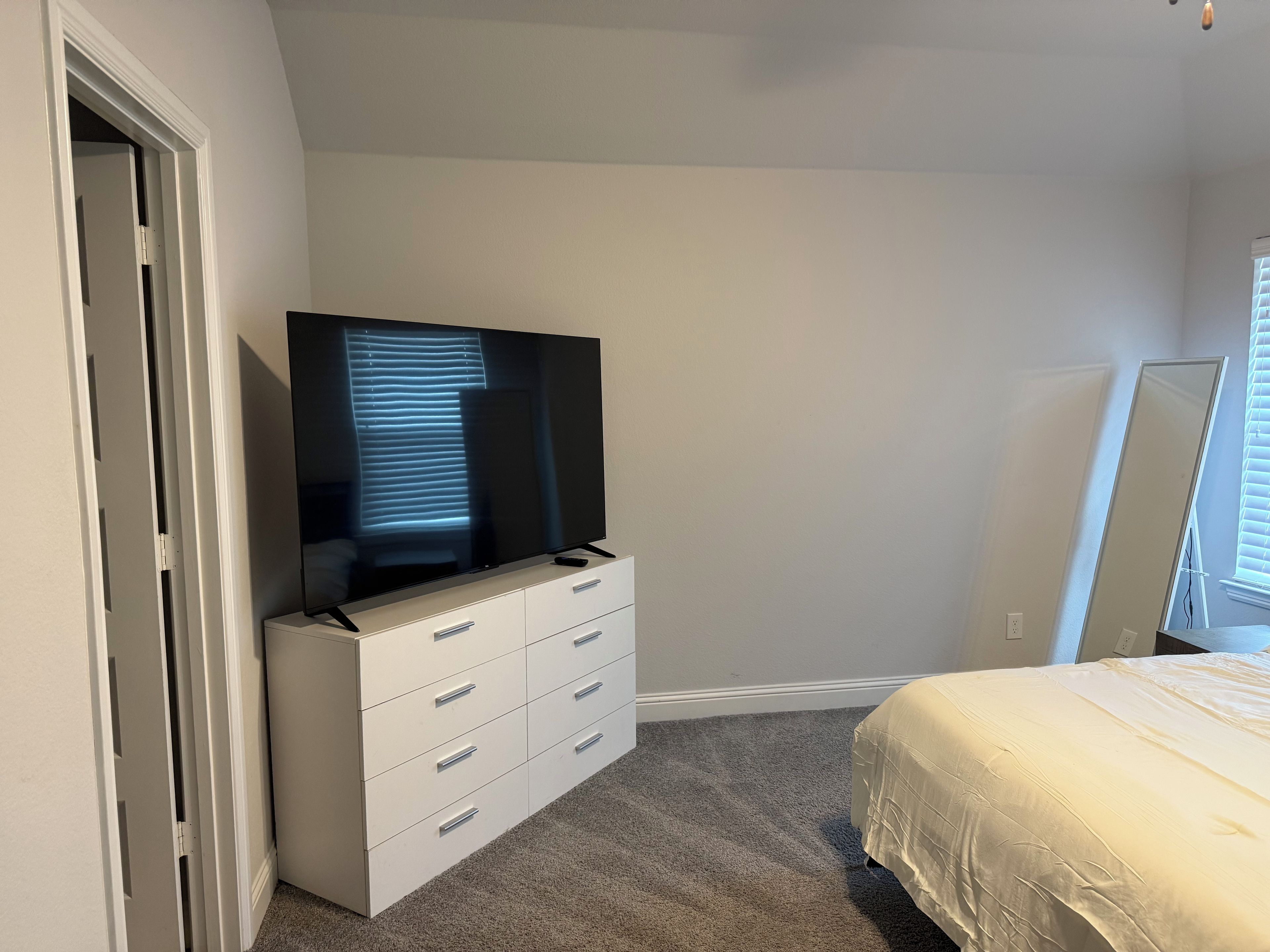 Ein Schlafzimmer mit Fernseher auf einer Kommode, einem Bett mit weißer Bettdecke und einem Fenster mit Jalousien.