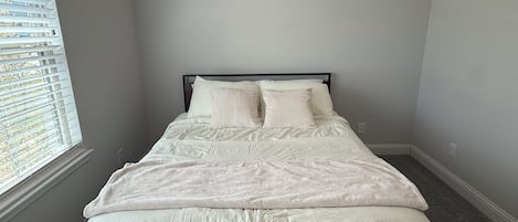 Un dormitorio con cama, ropa de cama blanca, una ventana con persianas y piso alfombrado.