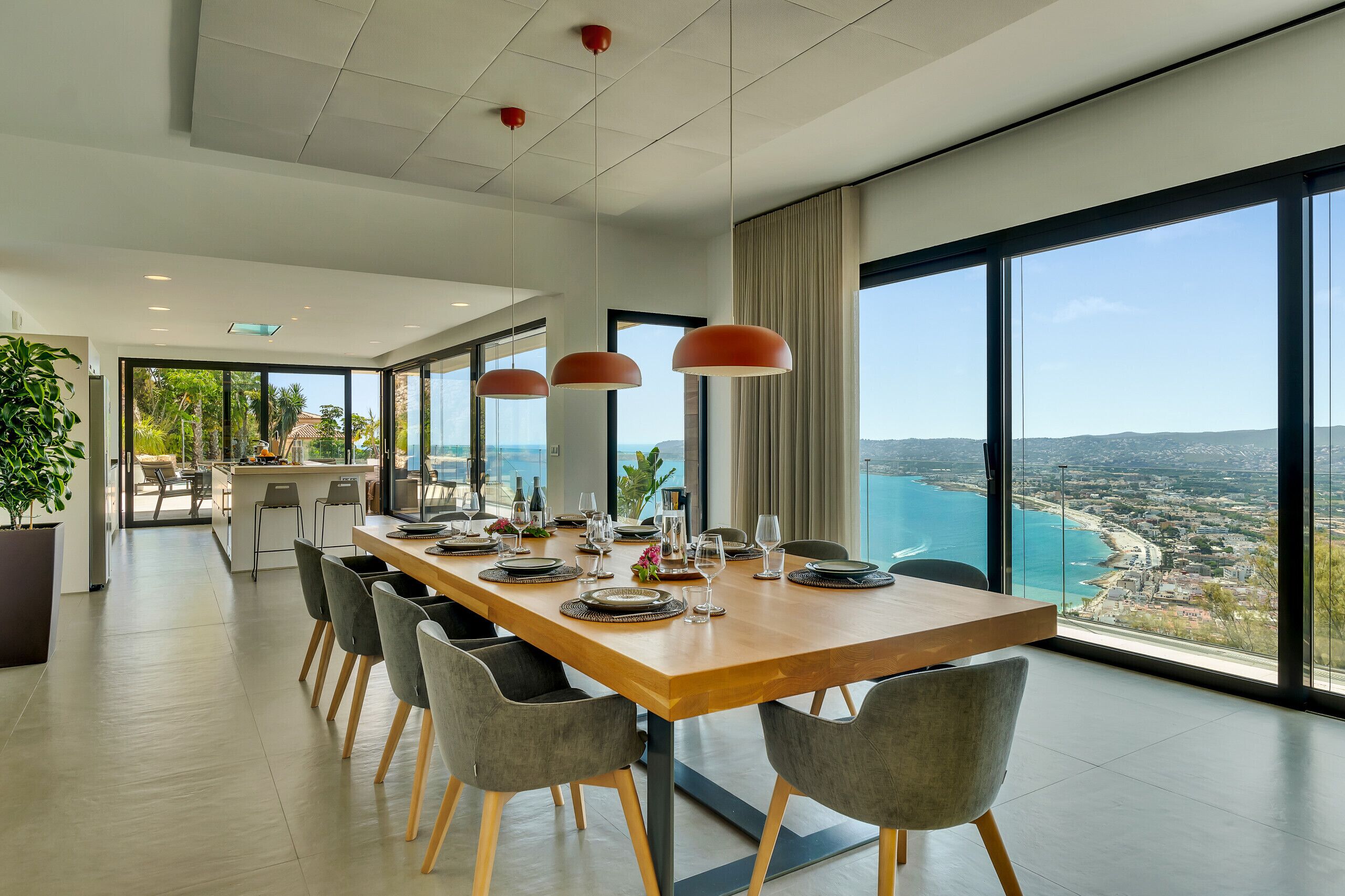 A modern dining area with a long table set for a meal, large windows offering a view of the sea and cityscape, and a kitchen area visible in the background.