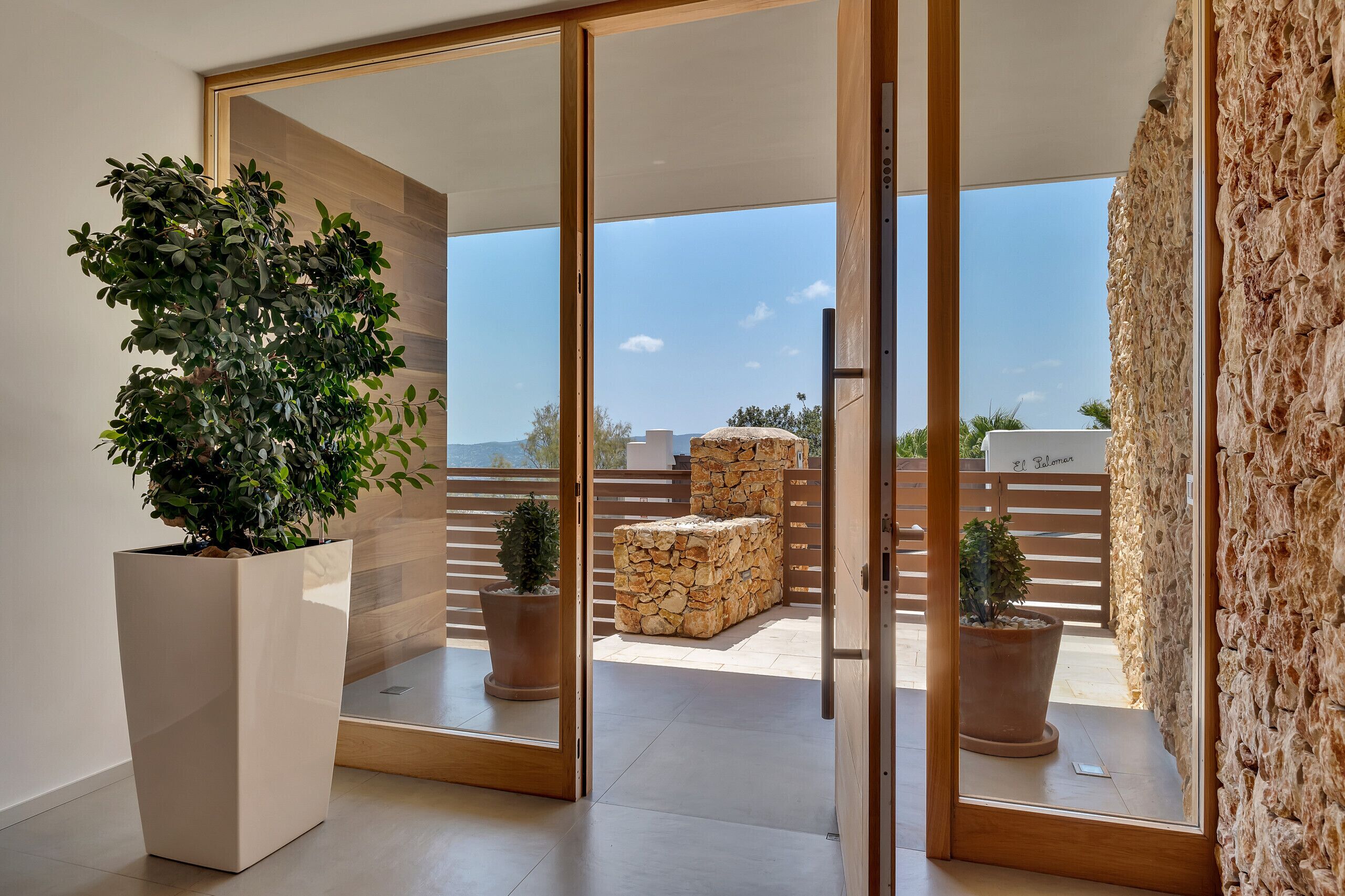 A modern interior with a large potted plant, a stone wall, and a view of a balcony with outdoor seating.