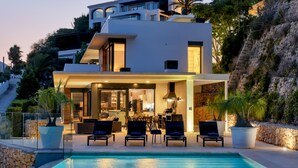 Ett modernt hus i två våningar med en swimmingpool, en utomhusplats med sittmöbler och utsikt över en bergig sluttning.