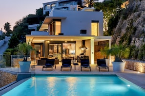 Ein modernes, zweistöckiges Haus mit Swimmingpool, Sitzgelegenheit im Freien und Blick auf einen felsigen Hang.