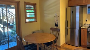 A kitchen with a stainless steel refrigerator, wooden cabinets, a round dining table with chairs, and a window with a view of a deck and greenery. - Urban Refuge in a welcoming East Bay  Neighborhood (Oakland)