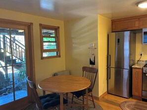 A kitchen with a stainless steel refrigerator, wooden cabinets, a round dining table with chairs, and a window with a view of a deck and greenery. - Urban Refuge in a welcoming East Bay  Neighborhood (Oakland)