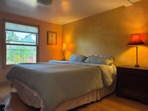 A bedroom with a bed, a nightstand, a lamp, a window with a view of greenery, and a framed picture on the wall. - Urban Refuge in a welcoming East Bay  Neighborhood (Oakland)