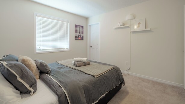 A bedroom with a bed, a window with blinds, a door, and wall-mounted shelves with decorative items. - Luxury 4BR Getaway|Mins to Falls & Casino|Sleeps 9 (Niagara Falls)