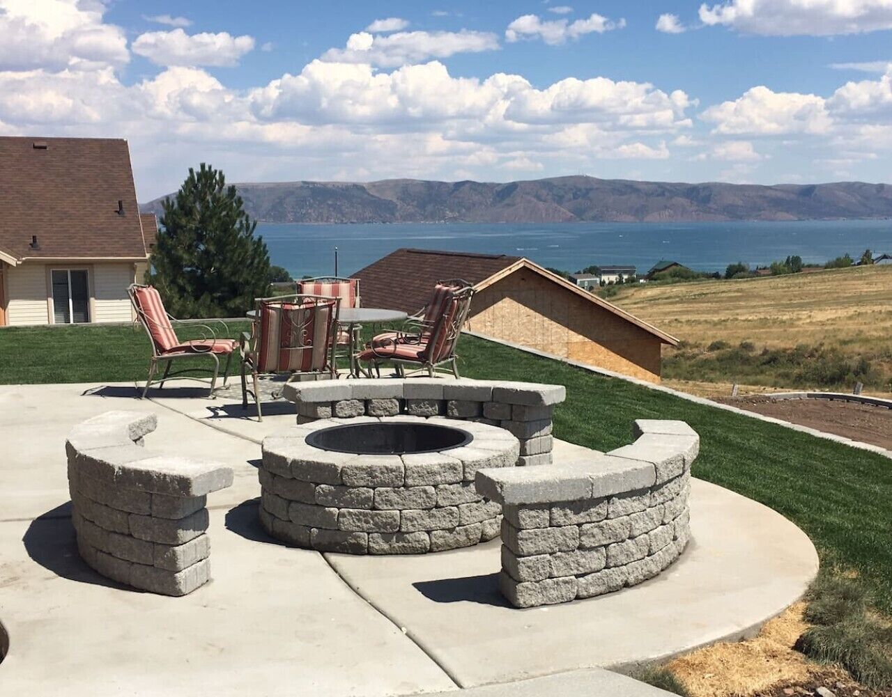 A backyard with a stone fire pit, outdoor furniture, and a view of a lake and hills.