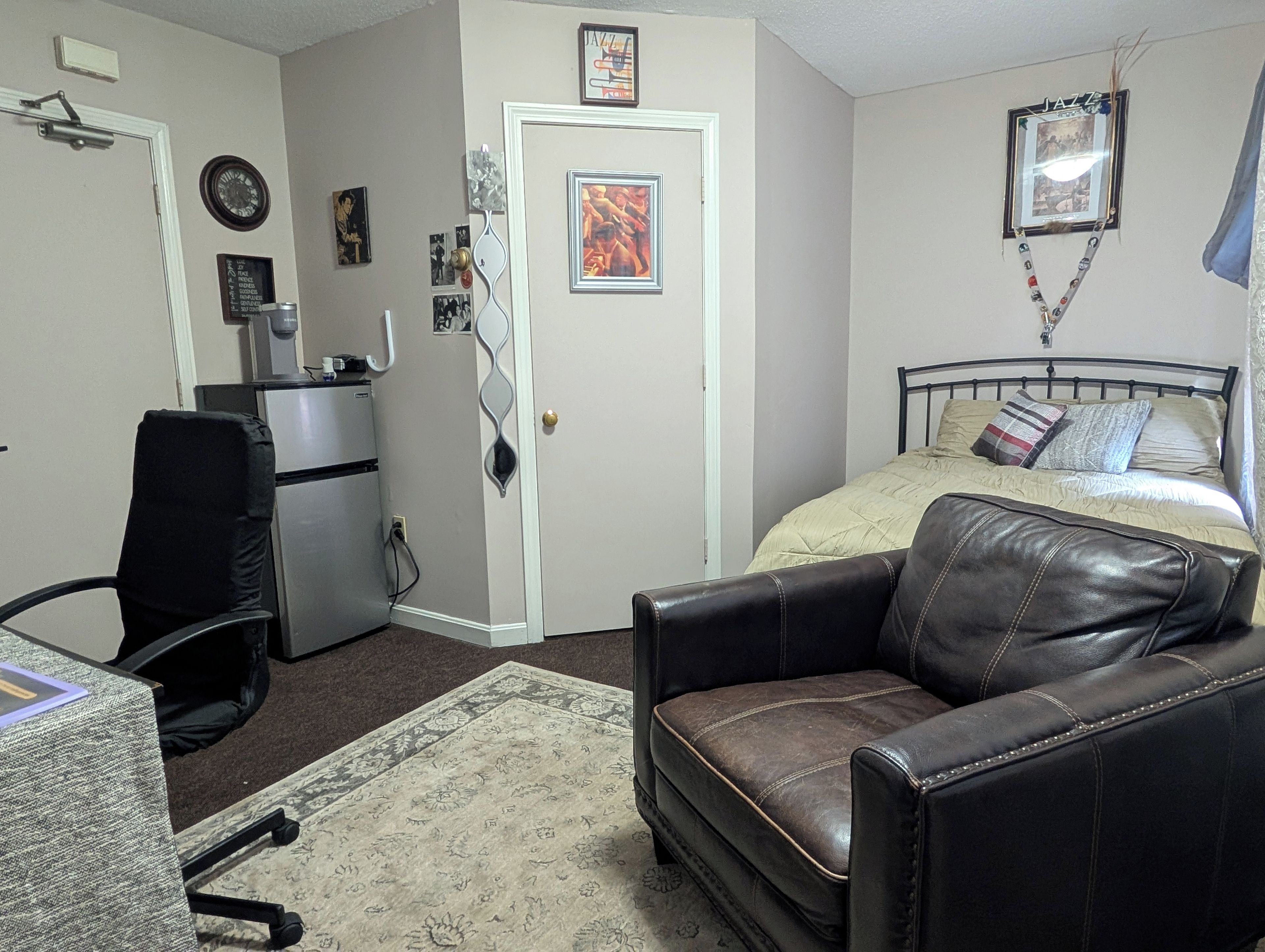 A bedroom with a bed, a desk, a chair, and a refrigerator.