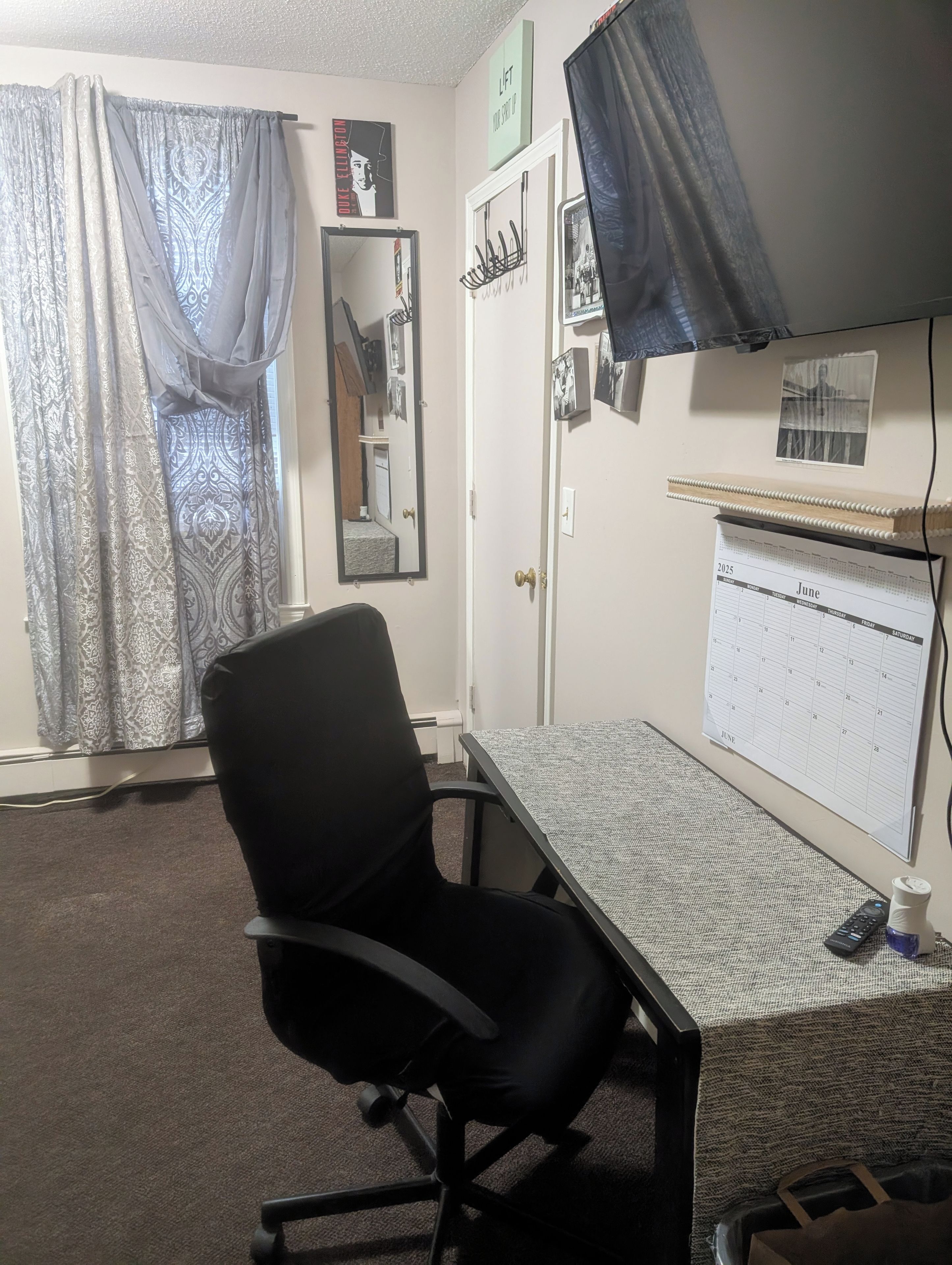 A room with a desk, chair, calendar, and curtains.